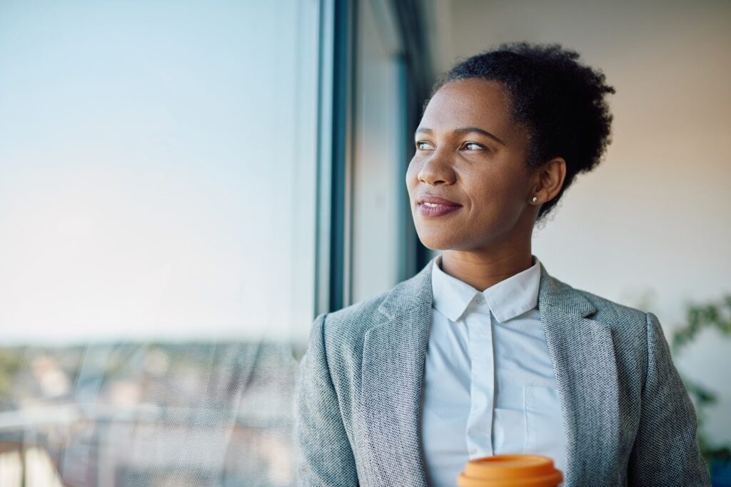 Close up of a woman in business attire holding a cup of coffee and staring out a large window.