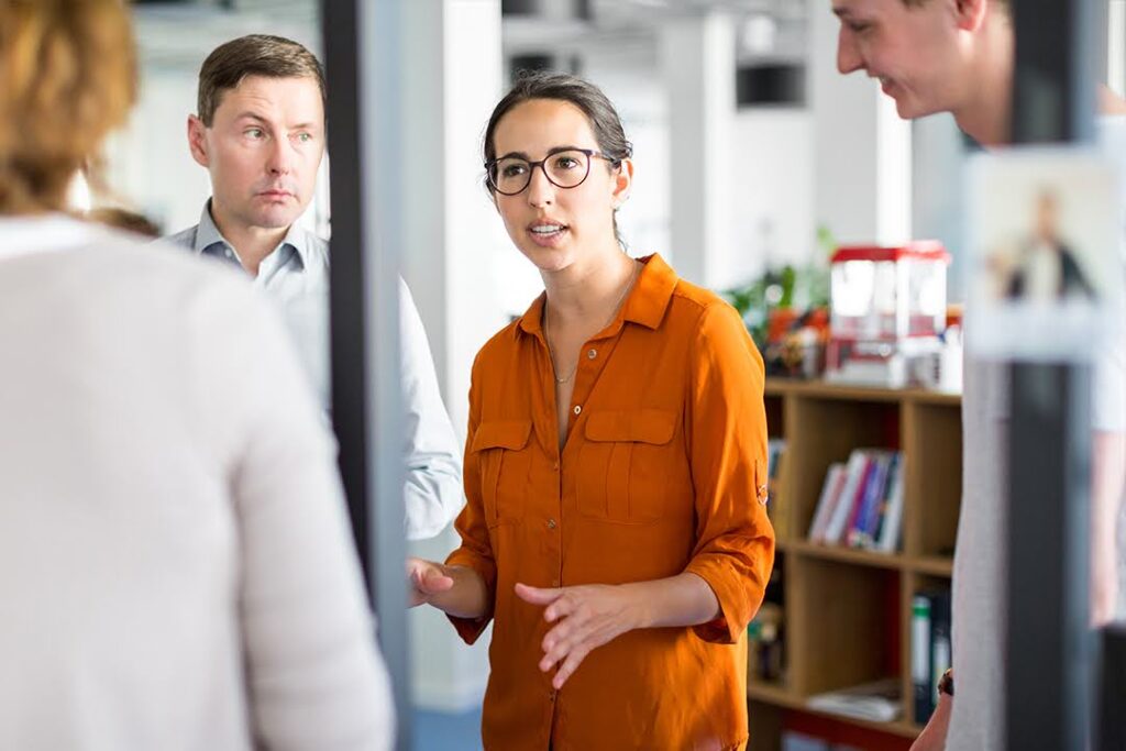 A woman in an orange shirt leads a discussion with colleagues in a modern office.