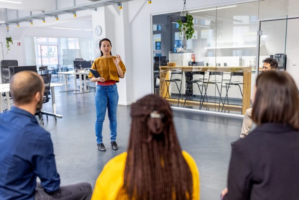 A woman stands and speaks to a seated group in a modern office space, holding a tablet and gesturing during a team meeting or presentation.